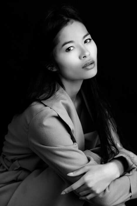 Portrait en noir et blanc d'une femme aux longs cheveux noirs, regardant pensivement l'appareil photo. Capturée lors d'une séance photo en studio à Lyon, elle est assise légèrement tournée, les bras croisés, vêtue d'un manteau clair sur un fond sombre.