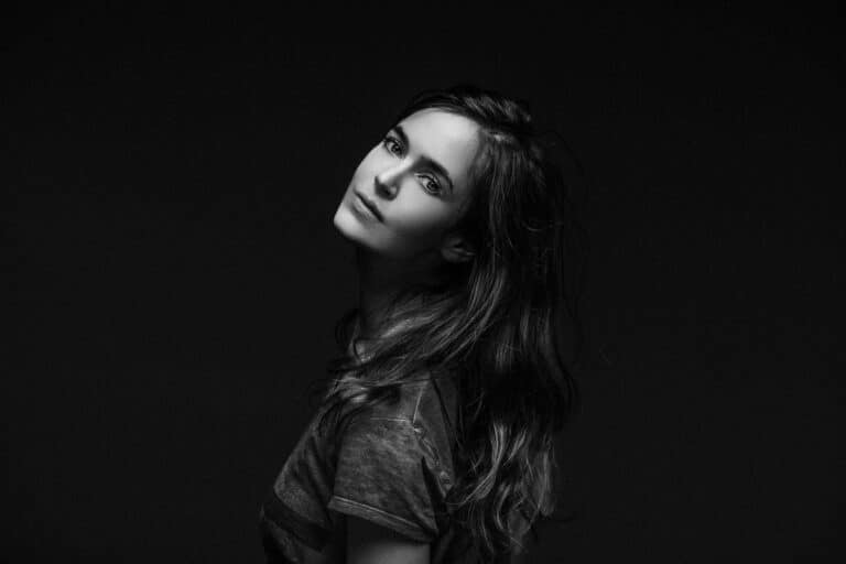 Une jeune femme aux cheveux longs et ondulés pose sur un fond sombre dans cette photo de studio à Lyon. Elle penche légèrement la tête et regarde doucement l'appareil photo. Le style noir et blanc met en valeur ses traits, en ajoutant un contraste et une texture spectaculaires.
