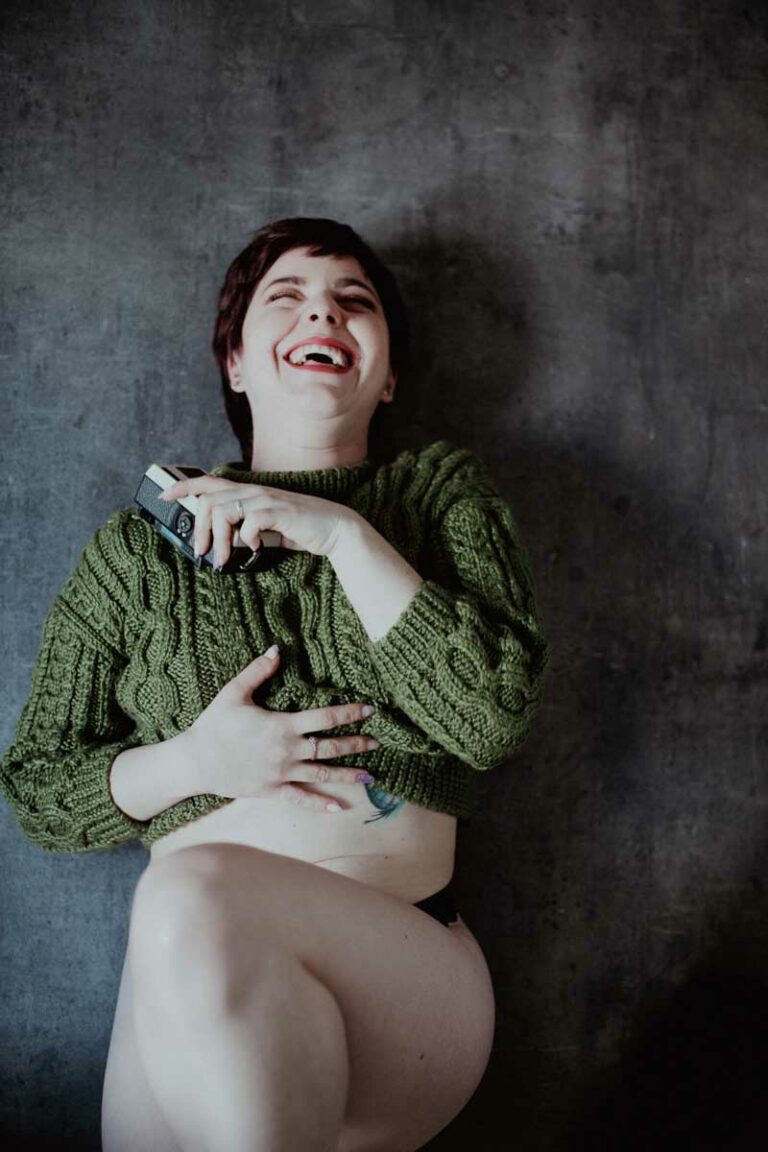 Une personne aux cheveux bruns courts, vêtue d'un gros pull vert et de sous-vêtements noirs, est assise contre un mur à la texture sombre. Elle rit joyeusement lors d'une photo de séance, les yeux fermés, tenant un objet rectangulaire et révélant un tatouage sur le ventre.