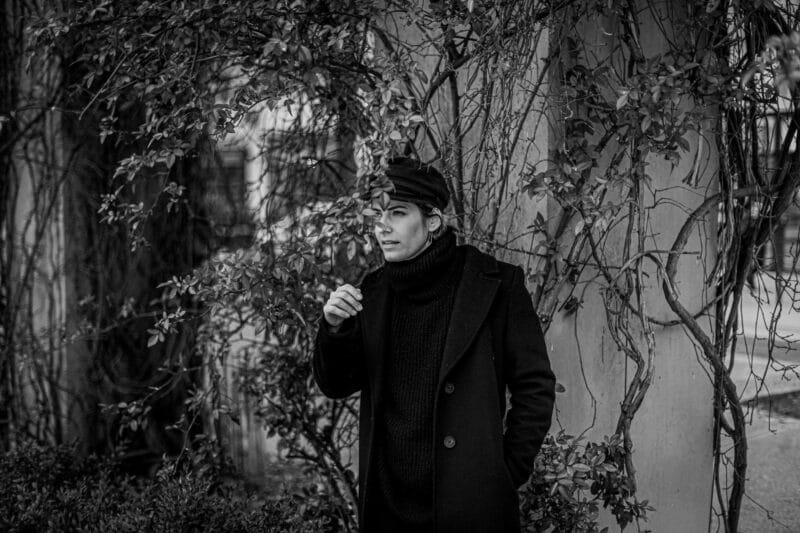 Une personne vêtue d'un manteau et d'un chapeau sombres se tient à côté d'un mur couvert de vignes, partiellement caché par des branches. Comme un mannequin perdu dans l'ado, elle a une main dans la poche et tient une cigarette, regardant pensivement sur le côté. L'image est en noir et blanc.