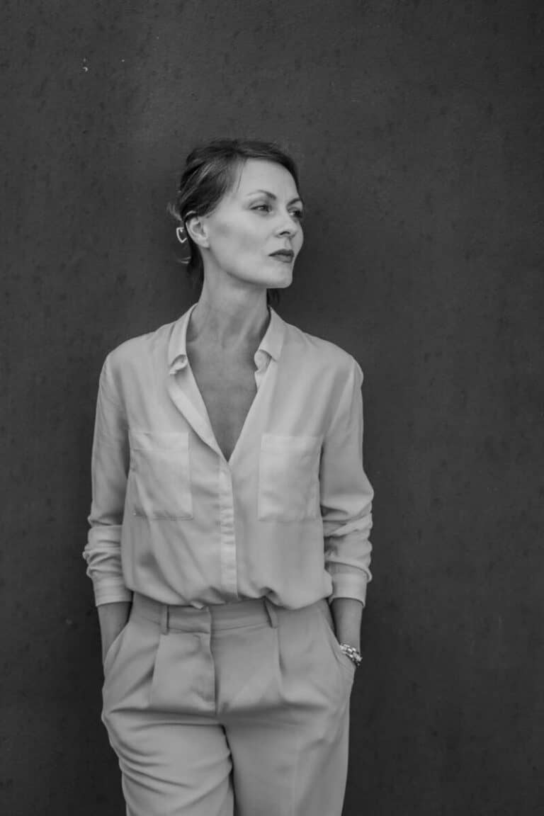 Photo en noir et blanc d'une femme debout contre un mur uni et sombre, réalisée pour une prise de vue par photographe macon. Elle porte une chemise à boutons rentrée dans un pantalon à taille haute, les mains dans les poches, regardant sur le côté avec une expression neutre.