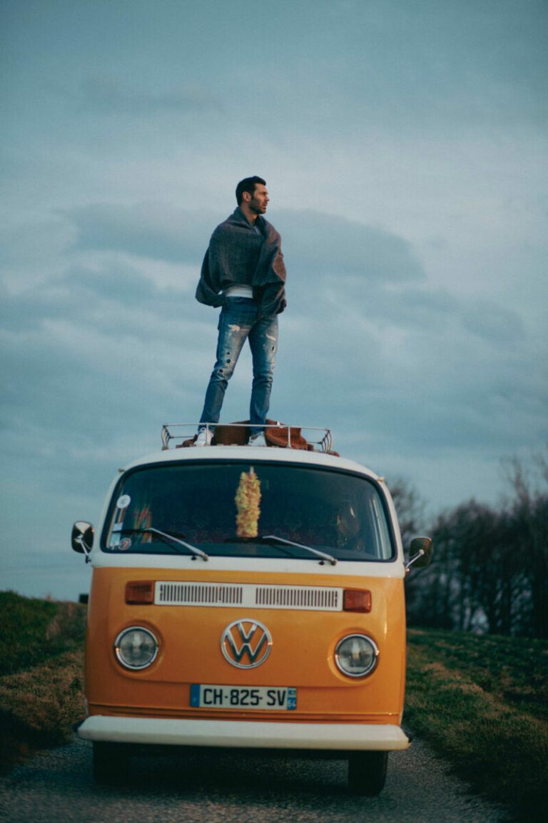 Un homme en jean et veste grise se tient sur le toit d'une camionnette Volkswagen jaune avec une plaque d'immatriculation française, garée sur une route de campagne. Cette photo de prise de vue capture le ciel nuageux et les arbres en arrière-plan.