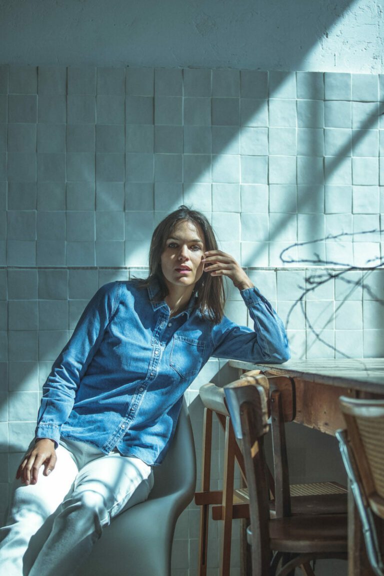 Une femme vêtue d'une chemise en jean bleu et d'un pantalon blanc est assise sur une chaise près d'une table en bois, la lumière du soleil entrant par la fenêtre. Capturée lors d'une prise de vue en studio photo Lyon, la scène respire le calme et la contemplation avec des ombres douces derrière elle.