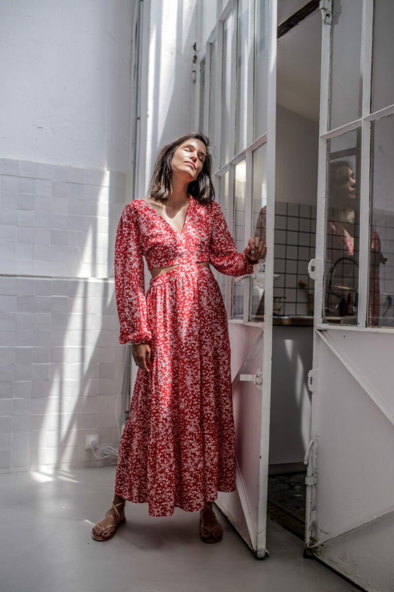 Une femme vêtue d'une longue robe rouge à fleurs se tient dans l'embrasure d'une porte éclairée par le soleil lors d'une séance photo en studio à Lyon, les yeux fermés et la tête légèrement inclinée vers l'arrière. Elle a des cheveux foncés et raides et des sandales brunes. La lumière du soleil projette des ombres sur les murs blancs derrière elle.
