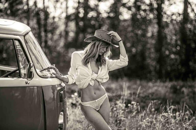 Une femme portant une chemise blanche nouée, des sous-vêtements en dentelle blanche et un chapeau de cow-boy s'appuie sur une camionnette d'époque dans un cadre extérieur ensoleillé et herbeux. Capturée en noir et blanc par un photographe pro sur Lyon, les arbres sont flous à l'arrière-plan.