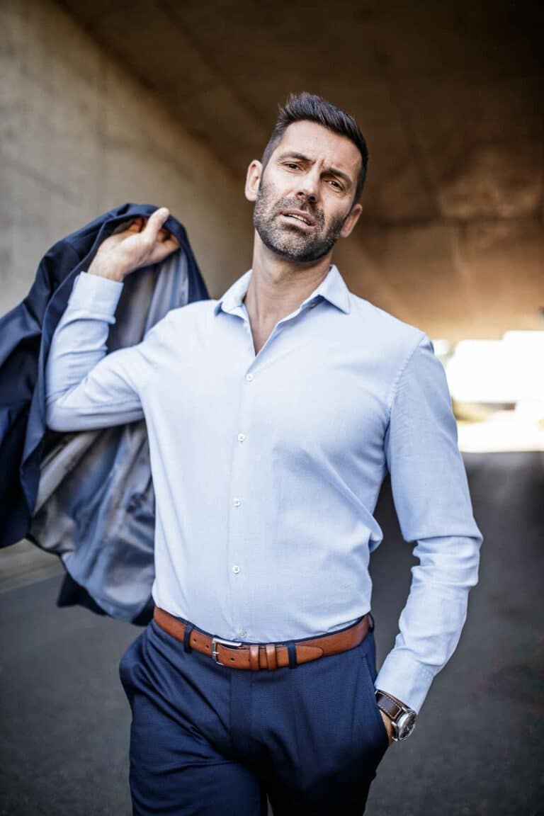 Un homme aux cheveux bruns courts et à la barbe porte une chemise bleu clair et un pantalon bleu marine avec une ceinture marron. Il tient avec assurance une veste de costume bleu marine sur son épaule lors d'un shooting photo à Lyon, debout sous un viaduc avec la lumière du soleil en arrière-plan.
