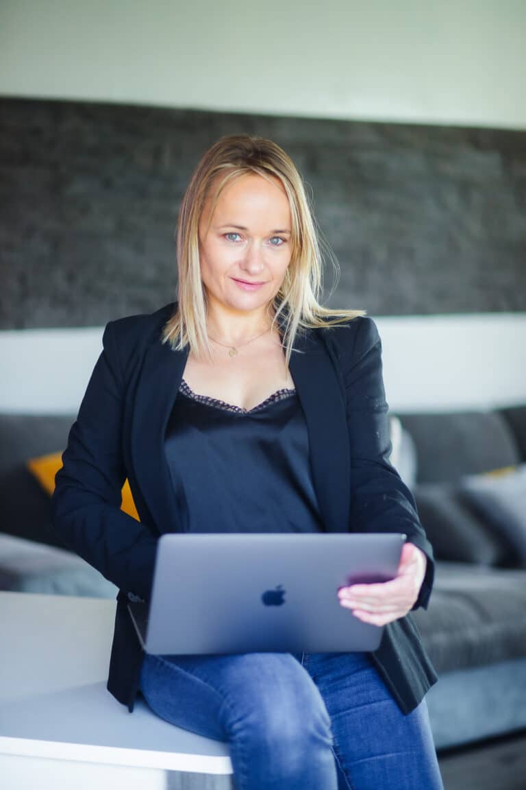 Une femme aux cheveux blonds et raides, vêtue d'un blazer noir et d'un haut sombre, est assise sur une table blanche et tient un MacBook ouvert. Capturée lors d'une séance photo par un photographe pro sur Lyon, elle sourit légèrement devant un canapé moderne gris avec des coussins jaunes.