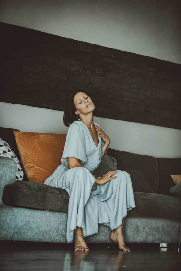 Une femme vêtue d'une robe fluide bleu clair est assise pieds nus sur un canapé gris avec des coussins orange et à motifs. Elle ferme les yeux, détendue et sereine, lors d'une prise de vue réalisée par un photographe pro sur Lyon dans un salon moderne à la lumière tamisée.