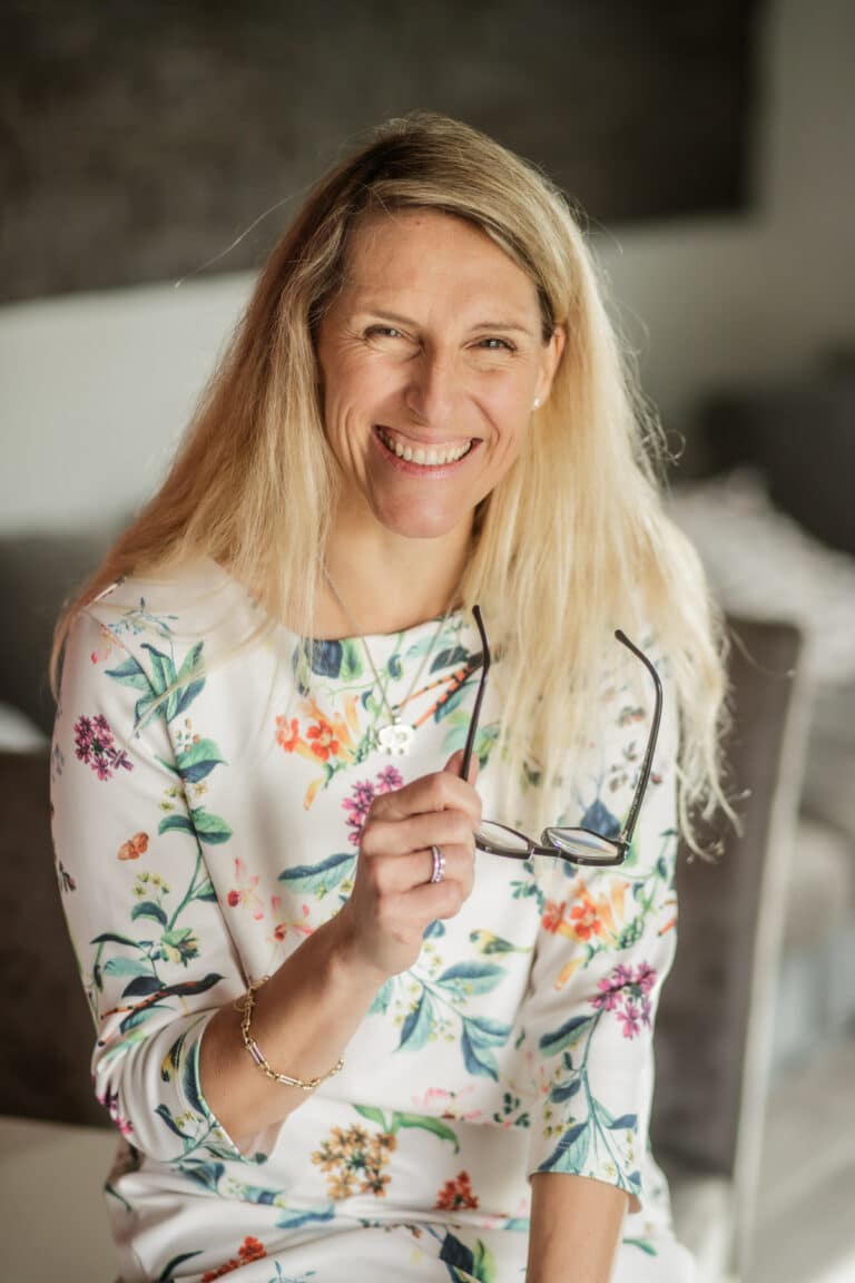 Une femme souriante aux longs cheveux blonds est assise à l'intérieur et tient des lunettes dans une main. Elle porte une robe blanche à motifs floraux colorés et un bracelet, dans ce qui semble être une photo de studio à Lyon, avec des meubles flous aux tons neutres derrière elle.