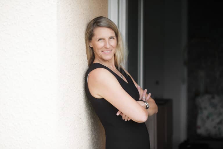 Une femme aux longs cheveux blonds et vêtue d'une robe noire sans manches se tient à l'intérieur de Lyon, adossée à un mur clair, les bras croisés. Elle sourit doucement à l'appareil photo lors d'une prise de vue faiblement éclairée, avec de légers meubles visibles à l'arrière-plan.