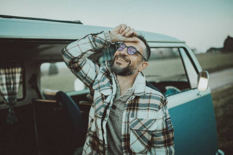 Un homme souriant, barbu et portant des lunettes, se tient à côté de la porte ouverte d'une camionnette bleue lors d'une prise de vue. Il porte une veste à carreaux et porte une main à son front. L'arrière-plan est herbeux, avec des rideaux visibles à l'intérieur de la camionnette.