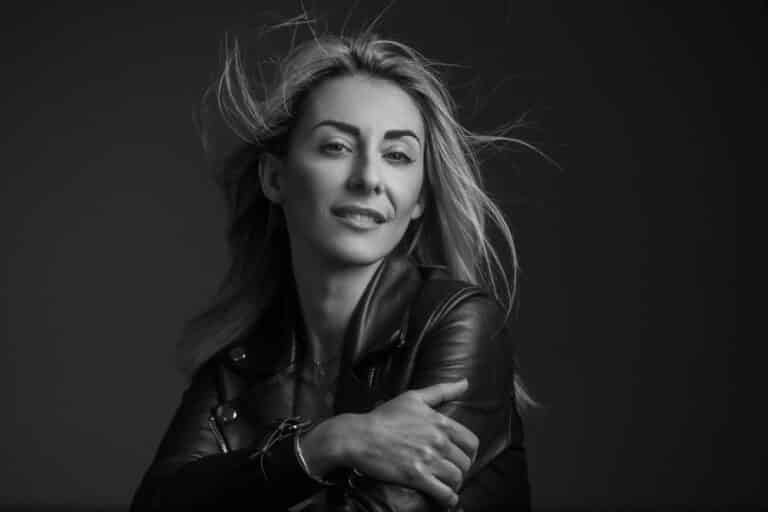 Portrait noir et blanc d'une femme aux cheveux longs, vêtue d'une veste en cuir. Elle se tient sur un fond sombre, avec un léger sourire sur le visage, regardant la caméra avec confiance. Ses cheveux flottent doucement au vent.