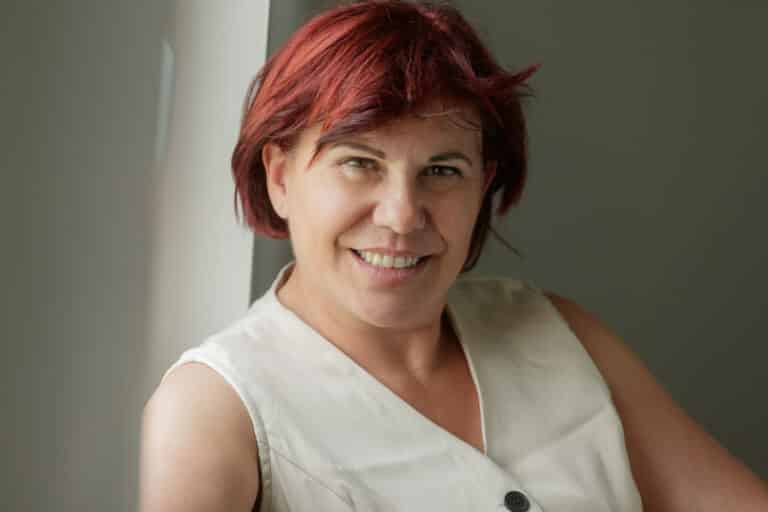 Une personne aux cheveux courts et roux et un haut sans manches de couleur claire sourit en s'appuyant contre un mur de couleur claire. L'individu a une expression détendue et amicale. L’arrière-plan est simple et neutre, gardant l’accent sur la personne.