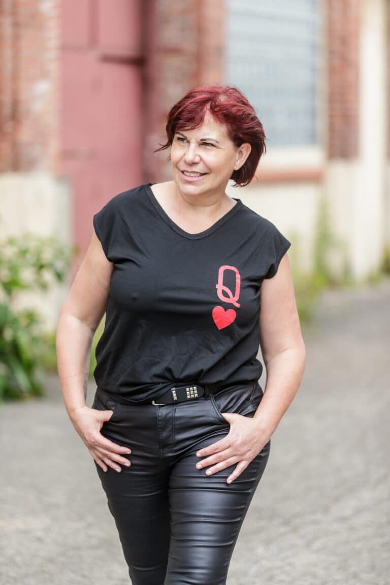 Une femme aux cheveux courts brun rougeâtre portant un t-shirt noir avec un cœur rouge et la lettre « Q » blanche sur le devant marche dehors. Elle porte également un pantalon en cuir noir et a les mains dans les poches. L’arrière-plan présente un bâtiment en brique avec une grande fenêtre et de la verdure.
