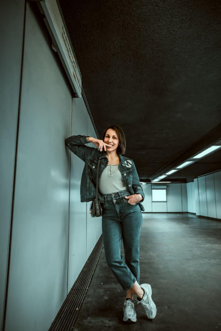 Une femme en tenue décontractée se tient dans un couloir aux murs gris et au plafond sombre, qui rappelle celui que l'on pourrait trouver lors d'un photographe pour shooting photo dans le métro de Lyon. Elle porte une veste en jean, un haut blanc, un jean taille haute et des baskets blanches. Elle s'appuie contre le mur avec un bras, pose son autre main sur son visage et sourit. Le couloir est vide