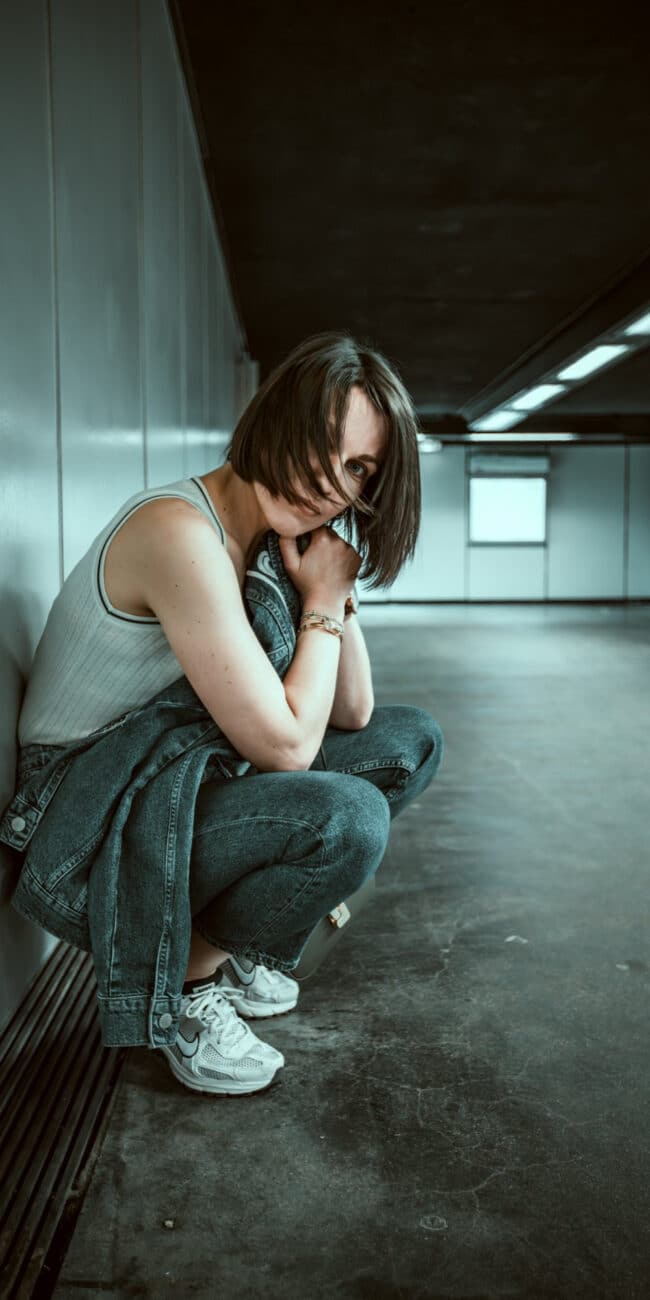 Une personne aux cheveux courts et foncés est accroupie dans un couloir étroit et faiblement éclairé avec des murs blancs et un sol sombre. Ils portent un débardeur blanc, un jean bleu et des baskets blanches. Tenant une veste en jean et posant leur menton sur leur main avec une expression pensive, ils ont l'air d'attendre qu'un photographe prenne une photo dans le métro de Lyon.