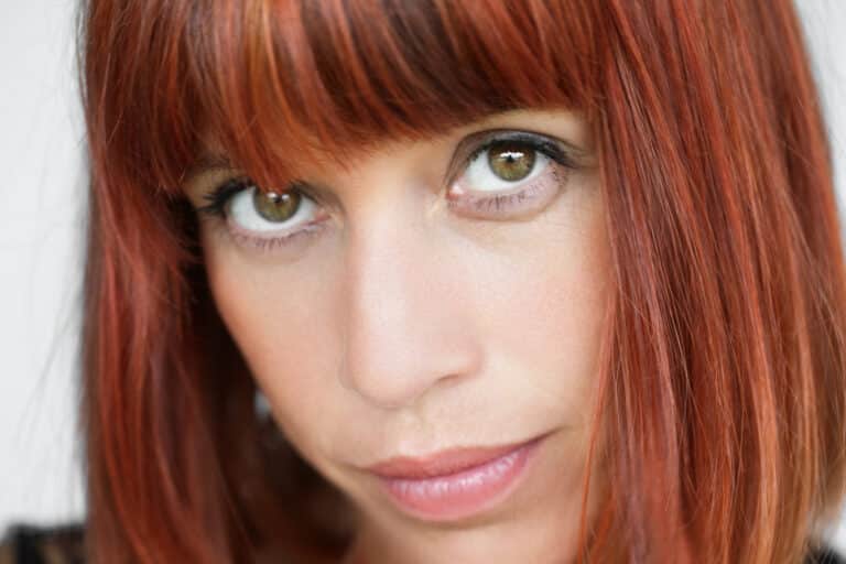 Portrait en gros plan d'une personne aux cheveux roux droits et mi-longs et à la frange. L'individu a la peau claire, les yeux verts et une expression faciale neutre, avec une tête légèrement inclinée et un regard direct vers la caméra. Parfait pour tout livre de photographe mettant en valeur le talent comédien de Lyon, l'arrière-plan est flou et neutre.