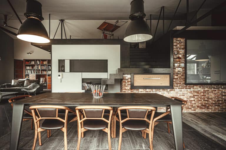 Espace de vie moderne de style industriel avec un coin repas au premier plan comprenant une table sombre, des chaises en bois et un verre de crayons. Derrière, il y a un escalier menant à un loft avec des murs en briques apparentes, des poutres sombres, de grandes suspensions et un coin salon confortable avec des étagères d'un côté, parfait pour être photographié par n'importe quel photographe lyonnais.
