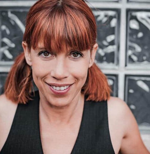 Une femme aux cheveux rouges coiffés de nattes sourit à la caméra. Elle porte un haut sans manches noir et se tient devant un fond de briques de verre texturées.