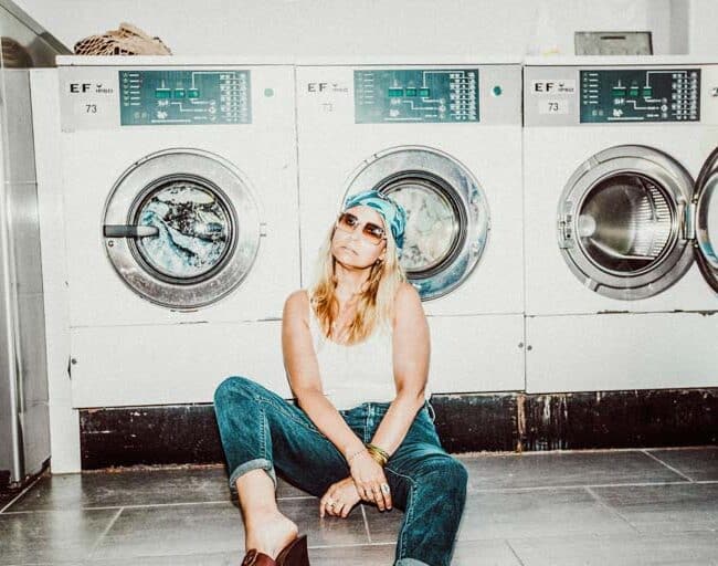 Une femme aux cheveux longs, portant des lunettes de soleil et un foulard, est assise par terre devant les machines à laver dans une laverie automatique. Elle est vêtue d'un jean et d'un débardeur blanc, penchée en arrière, les jambes tendues dans une pose détendue.