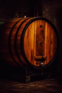 Un grand tonneau de vin en bois se dresse debout sur un solide cadre noir dans une pièce faiblement éclairée, presque comme s'il posait pour un photographe bouteille de vin. Le fût est fait d'un bois riche et brun doré entouré de bandes de métal foncé. Un robinet en argent est fixé près du bas, avec une petite chaîne qui pend. L’arrière-plan est un mur sombre et texturé.