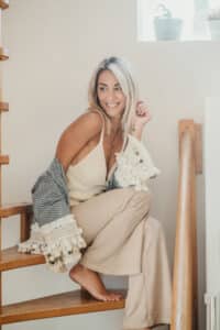 Une femme blonde au sourire enjoué est assise sur un escalier en bois et prend des photos dans sa charmante demeure de Lyon. Elle est vêtue d'un débardeur beige, d'un pantalon marron clair et d'un châle rayé à pompons. Une petite plante en pot repose sur le rebord de la fenêtre derrière elle et la lumière naturelle inonde l'espace.