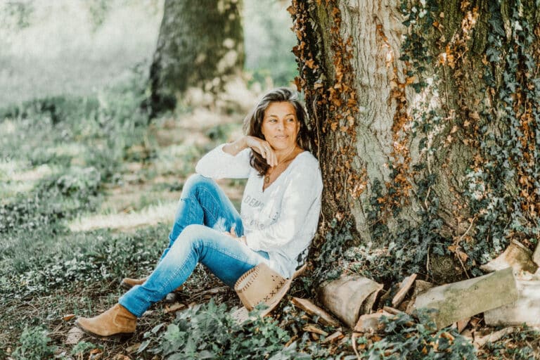 Dans un cadre extérieur paisible, une femme est assise contre un tronc d'arbre lors d'une séance photo à Lyon. Elle porte un pull léger, un jean et des bottes marron. Son regard dérive vers le côté au milieu des feuilles mortes et de la verdure, tandis que la lumière du soleil filtre à travers les arbres, projetant une lueur sereine.