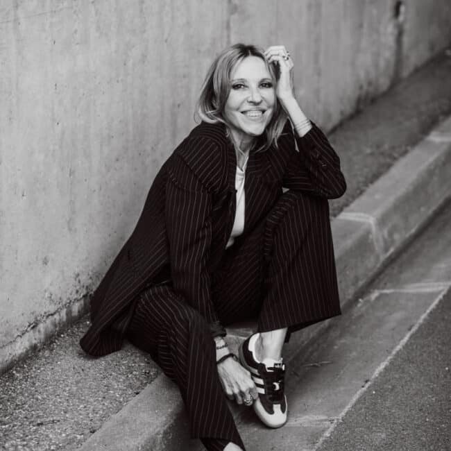 Une femme portant un costume à rayures et des baskets est assise sur un trottoir contre un mur en béton lors d'une séance photo à Lyon. Elle sourit, une main posée sur son genou et l'autre touchant ses cheveux. L'image en noir et blanc dégage une atmosphère décontractée et confiante.