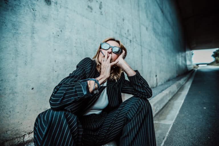 Un homme en costume rayé et lunettes de soleil est assis contre un mur de béton dans un tunnel, rappelant une séance photo à Lyon. Il se couvre la bouche avec ses deux mains, montrant divers bracelets. L'arrière-plan révèle les murs courbes du tunnel et un chemin menant à l'extérieur.