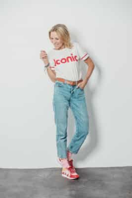 Une femme portant un t-shirt blanc avec l'inscription « iconique » en rouge, ainsi qu'un jean bleu, pose pour une séance photo à Lyon. Portant des baskets rouges et blanches avec des chaussettes roses, ses cheveux blonds mi-longs complètent son sourire éclatant alors qu'elle se tient debout contre un mur blanc uni, la main sur la hanche et faisant tournoyer ses cheveux de manière ludique.