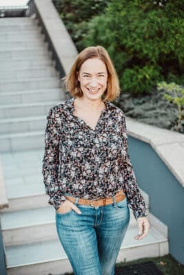Une femme aux cheveux mi-longs se tient à l'extérieur dans une séance photo décontractée à Lyon, souriante. Elle porte un chemisier noir à fleurs et un jean bleu, avec une ceinture marron. Ses mains reposent dans ses poches tandis qu'un escalier et de la verdure forment l'arrière-plan, renforçant l'atmosphère décontractée.