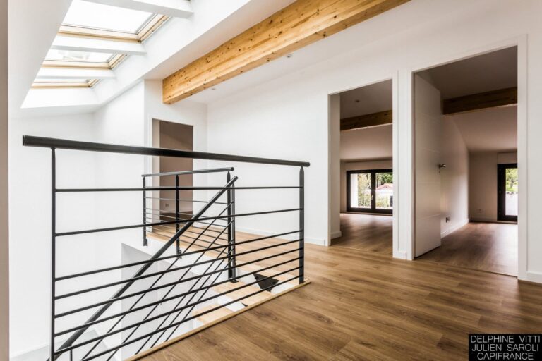 L'intérieur moderne aux murs blancs, au parquet et aux poutres apparentes au plafond reflète un style architectural d'inspiration lyonnaise. Des puits de lumière illuminent le palier à l'étage tandis qu'une rampe en métal noir entoure l'escalier. Les portes ouvertes révèlent des pièces adjacentes avec de grandes fenêtres et une végétation luxuriante à l'extérieur.