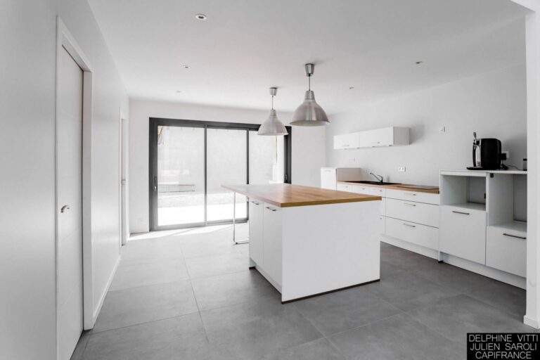 Cuisine moderne avec des armoires blanches et des comptoirs en bois, magnifiquement photographiées dans le cadre d'une architecture contemporaine. Un grand îlot est doté de deux luminaires suspendus au-dessus, tandis que des appareils électroménagers en acier inoxydable ornent la partie droite. Le sol carrelé gris et les portes coulissantes en verre offrent une lumière naturelle pour un design minimaliste.