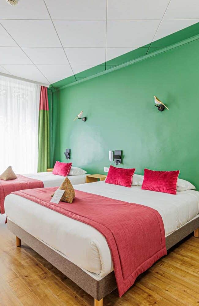 Une chambre d'hôtel lumineuse, idéale pour un photographe à Lyon, dispose d'un lit double et d'un lit simple avec une literie blanche impeccable, des touches de rouge et des appliques modernes. La lumière du soleil pénètre à flots par de grandes fenêtres ornées de rideaux verts et rouges, mettant en valeur l'élégant parquet.