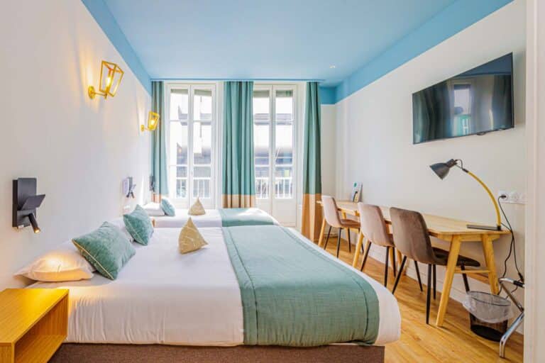 Chambre d'hôtel lumineuse avec deux lits doubles, chacun orné de plaids et de coussins verts. De grandes fenêtres avec des rideaux bleu sarcelle laissent entrer la lumière naturelle. Idéale pour un shooting photo à l'hôtel Lyon, cette chambre présente une décoration moderne, du parquet et des murs bleu-blanc.