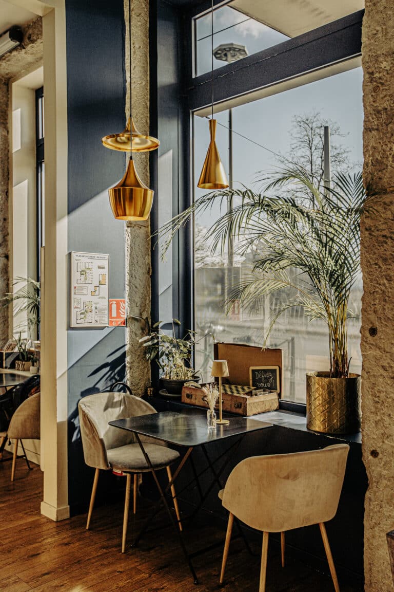 Un coin de café moderne et ensoleillé avec des murs bleus, des lampes suspendues dorées, des plantes en pot et des chaises beiges confortables - un endroit parfait pour un shooting photo à Lyon. De grandes fenêtres éclairent les planchers en bois, créant une atmosphère détendue et élégante.
