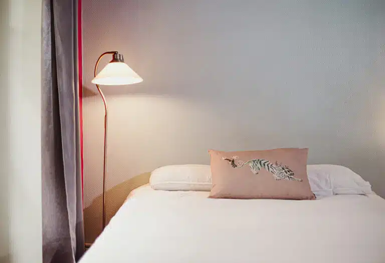 Un lit soigneusement fait avec des draps blancs, deux oreillers et un coussin décoratif rose clair orné d'un tigre brodé. Une lampe de chevet murale éclaire doucement, et un rideau gris pend à gauche, contre un mur clair texturé.