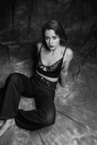 Une femme est assise sur un sol drapé de tissu, une jambe tendue et l'autre pliée. Elle porte une brassière en dentelle noire et un jean foncé. L'image est en noir et blanc, ce qui lui confère une atmosphère sombre et artistique. Elle regarde droit vers l'objectif avec une expression neutre.