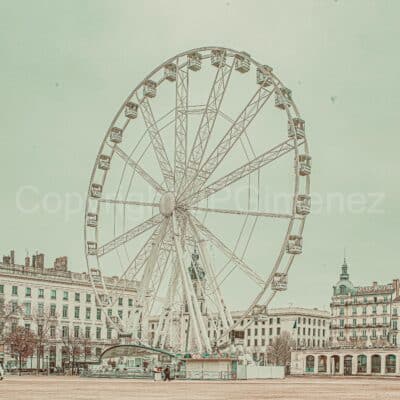 Une grande roue blanche trône sur une place ouverte, entourée de bâtiments historiques. Le ciel est pâle et couvert. Cette scène capture le charme des photographies lyonnaises, avec l'Hôtel Royal et l'architecture européenne classique en arrière-plan.