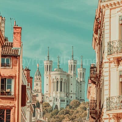 Une grande basilique blanche avec quatre tours et des détails ornés s'élève au-dessus des arbres, encadrée par de vieux bâtiments avec des balcons sous un ciel turquoise clair - un paysage urbain pittoresque et vintage vu dans les photographies lyonnaises.