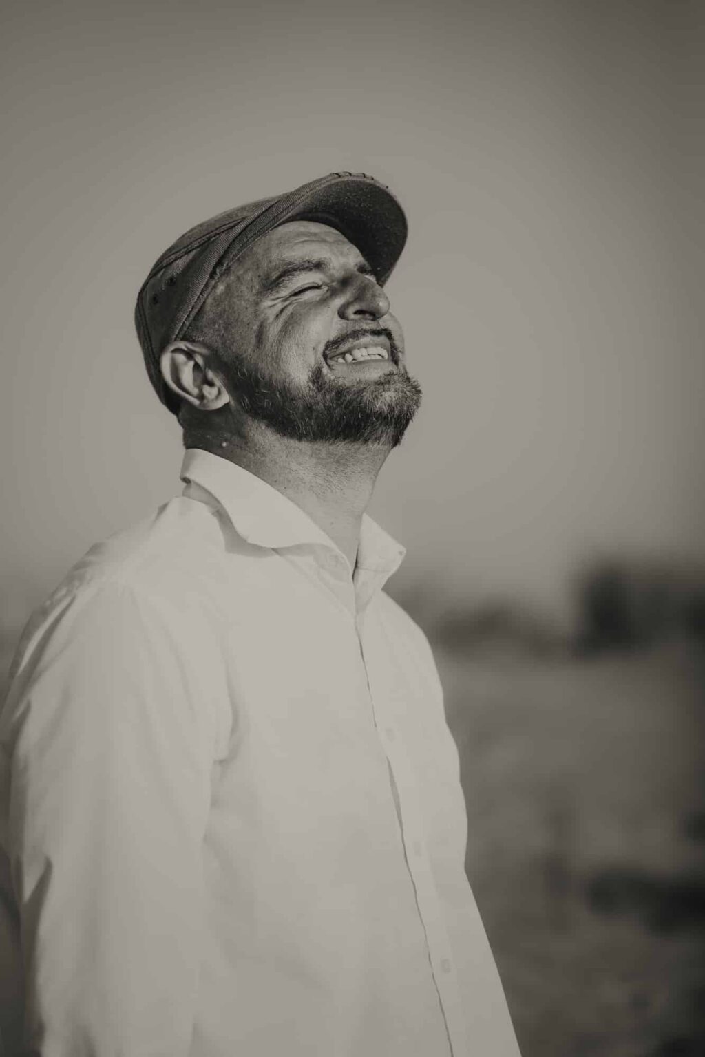 Un homme barbu, vêtu d'une chemise blanche et d'une casquette plate, sourit largement, les yeux fermés, en penchant la tête en arrière. La photo est en noir et blanc et l'arrière-plan est flou, ce qui crée une ambiance sereine et joyeuse.