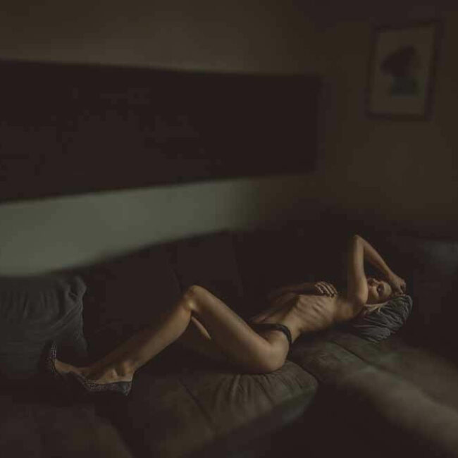 Une femme portant de la lingerie noire est allongée sur un canapé sectionnel sombre dans une pièce faiblement éclairée. Elle est allongée sur le dos, un bras au-dessus de sa tête et l'autre main sur sa poitrine. La lumière d'une fenêtre éclaire une partie de son corps et la pièce semble intime et morose.