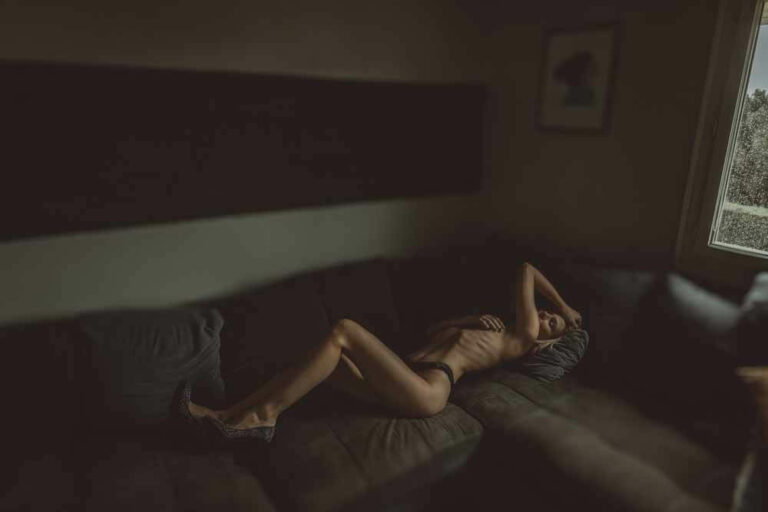 Une femme portant de la lingerie noire est allongée sur un canapé sectionnel sombre dans une pièce faiblement éclairée. Elle est allongée sur le dos, un bras au-dessus de sa tête et l'autre main sur sa poitrine. La lumière d'une fenêtre éclaire une partie de son corps et la pièce semble intime et morose.