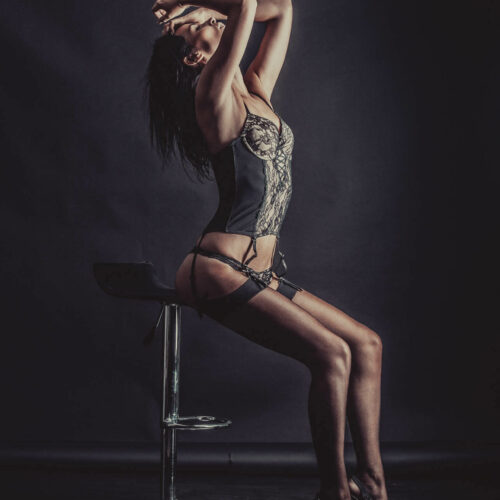 Une femme en lingerie, bas et talons hauts est assise sur un tabouret de bar moderne sur un fond sombre. Elle cambre son dos et étire ses bras au-dessus de sa tête, la tête inclinée vers l'arrière, créant ainsi une pose sensuelle. L'éclairage met en valeur sa silhouette et ses courbes.