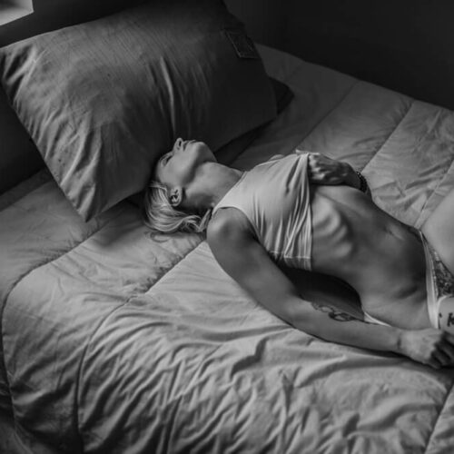Une personne aux cheveux courts est allongée sur un lit dans une pièce faiblement éclairée, vêtue d'un sous-vêtement et d'un haut court. Elle a une main sur la poitrine et l'autre sur le lit, la tête penchée en arrière. La photo est en noir et blanc, avec des ombres et des lumières douces.