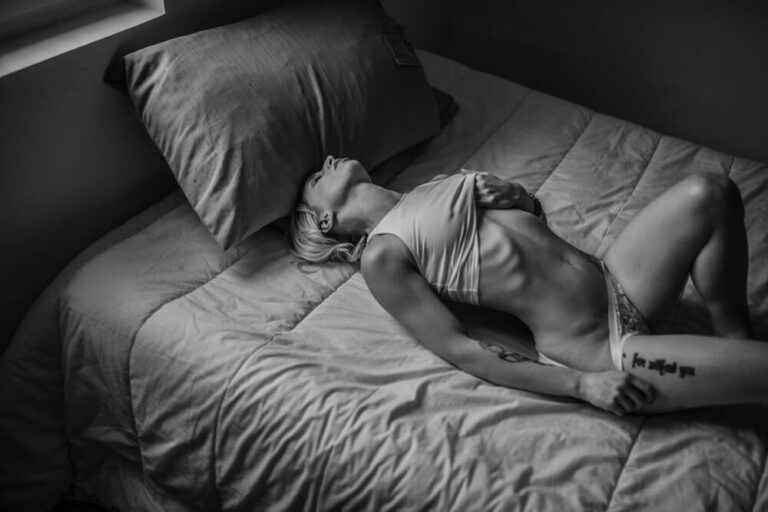 Une personne aux cheveux courts est allongée sur un lit dans une pièce faiblement éclairée, vêtue d'un sous-vêtement et d'un haut court. Elle a une main sur la poitrine et l'autre sur le lit, la tête penchée en arrière. La photo est en noir et blanc, avec des ombres et des lumières douces.