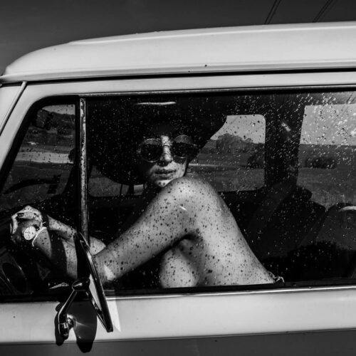 Une photo en noir et blanc montre une personne aux seins nus, portant des lunettes de soleil et un chapeau, assise sur le siège du conducteur d'un vieux véhicule. Son bras repose sur le cadre de la fenêtre et elle regarde en arrière par-dessus son épaule. La vitre est tachetée de gouttes d'eau.