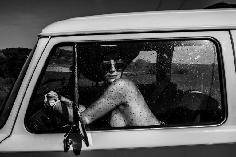 Une photo en noir et blanc montre une personne aux seins nus, portant des lunettes de soleil et un chapeau, assise sur le siège du conducteur d'un vieux véhicule. Son bras repose sur le cadre de la fenêtre et elle regarde en arrière par-dessus son épaule. La vitre est tachetée de gouttes d'eau.