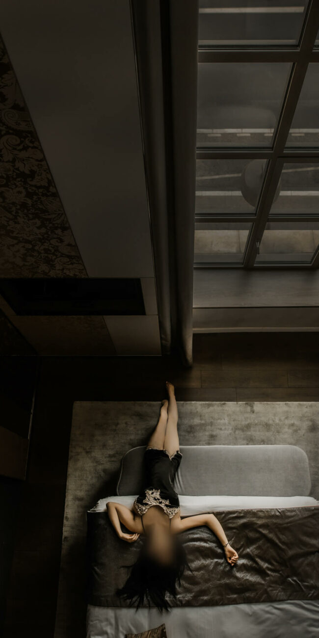 Une personne aux longs cheveux noirs est allongée sur le dos sur un lit dans une pièce moderne, faiblement éclairée, avec une grande fenêtre au-dessus de la tête. Elle porte un vêtement noir et a un bras tendu, tandis que ses jambes reposent nonchalamment sur le bord du lit.