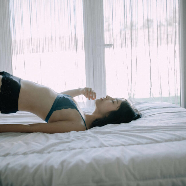 Une femme vêtue d'un soutien-gorge bleu et d'un short noir est allongée sur un lit blanc, le dos voûté et les jambes relevées. La lumière du jour traverse les rideaux transparents derrière elle, créant une atmosphère calme et sereine dans la chambre à coucher.